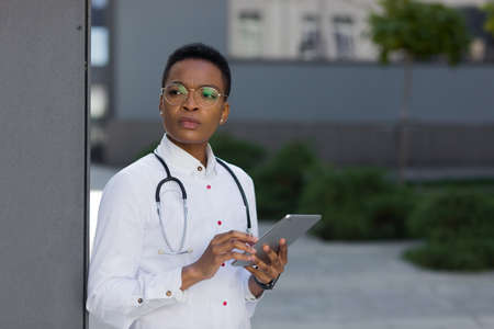 The doctor near the clinic holds a tablet computer, reads the news African-American seriously studies information from the Internet, for online consultations with patientsの写真素材