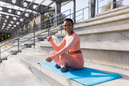 African American woman smiling and looking at camera, sitting in lotus position on fitness mat, confident and cheerful after yogaの写真素材