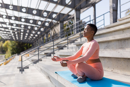 African American woman performs breathing exercises sitting on a yoga mat in the park near the stadium, resting after trainingの写真素材