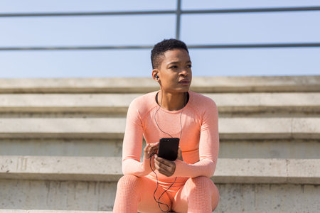 Afro american woman tired after workout listening to music and audio podcast, after jogging and...