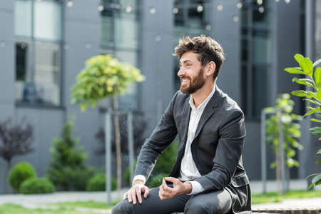 Caucasian bearded business man smoking cannabis outdoors sitting on city park bench on urban street background. Male employee Businessman. Office worker in suit Relieves stress a marijuana outsideの写真素材