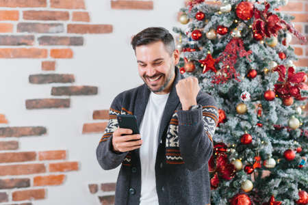 Young male freelancer businessman rejoices winning happy victory reads great news from mobile phone at home during New Year and Christmas holidaysの写真素材