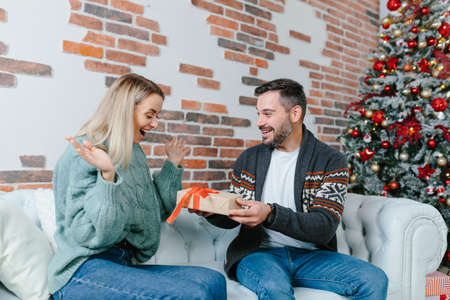 Young family together celebrate New Year and Christmas, couple man and woman give gifts to each other sitting on sofa at home happy and in loveの写真素材