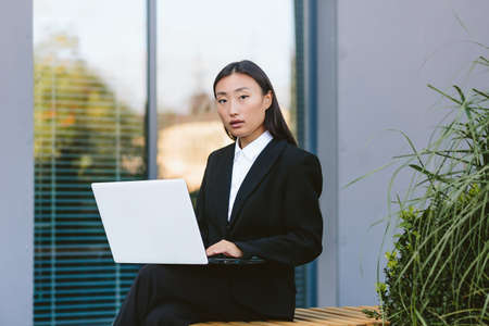 Successful asian woman businessman working on laptop outdoors sitting near office center and looking at cameraの写真素材