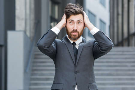 sad worried business bearded caucasian man holding his head. Businessman entrepreneur or employee or worker in suit on city street on background of modern office center. Outside. outdoorsの写真素材