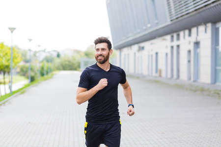 Young Caucasian bearded happy man runner jogging down the street on urban background of city park. Morning jog. Active healthy lifestyle fitness. Sport workout outdoor. Athlete training run exerciseの写真素材