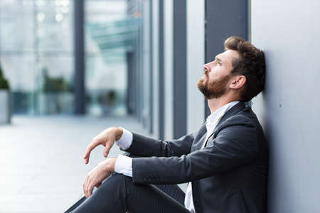Sad depressed entrepreneur in formal suit worker man sitting near outdoors street near modern office business center. Upset male businessman lost job due financial crisis employee has problem. outsideの写真素材