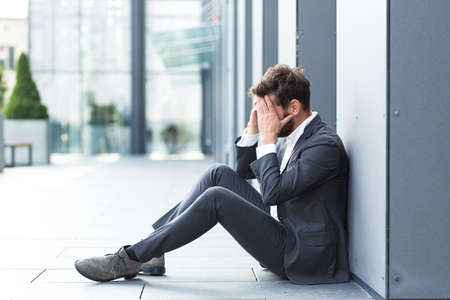 Sad depressed entrepreneur in formal suit worker man sitting near outdoors street near modern office business center. Upset male businessman lost job due financial crisis employee has problem. outsideの写真素材