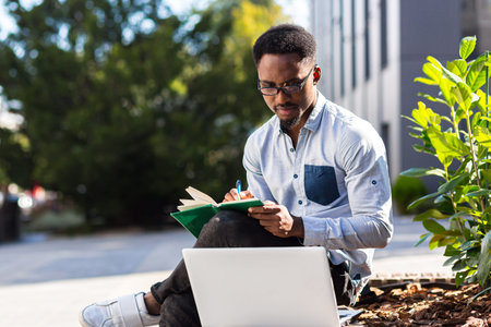 Young african american male student sitting in a city park on a bench with laptop and notebook...