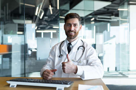 Successful doctor works sitting at the table looks at the camera and smiles makes a positive gesture with his hands happy, works in a modern clinicの写真素材