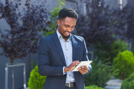 Young african american businessman in formal business suit standing with tablet in hands on...