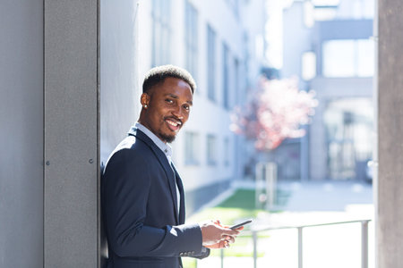 Successful and happy african american businessman near office holding mobile phone smiles and rejoices reads news and writesの写真素材