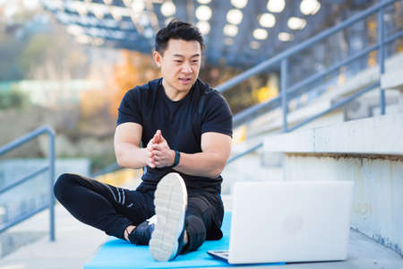 handsome asian fitness coach holds workout classes online using video call webcam and laptop. sitting outside in a modern stadium. Pilates training or stretching. Physical exercises. Records Lessonsの写真素材