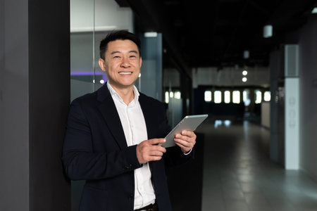 Successful Asian businessman boss man, in a business suit rejoices and smiles uses a tablet...
