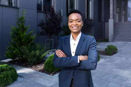Portrait of young successful african american business woman happy and smiling looking at camera with arms crossed, outside officeの写真素材