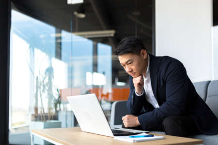 Portrait thoughtful Asian businessman working on a laptop computer at a modern office desk. Confident Focused pensive man in formal suit indoors. thinking of inspiration solving a problem. Startupの写真素材