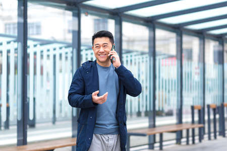 Cheerful and happy Asian businessman talking on the phone, waiting for a plane and a taxi at a public transport stopの写真素材