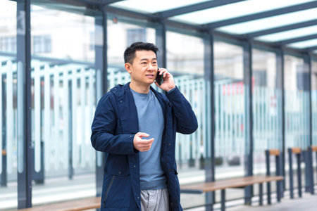 A man talking on the phone, focused and serious Asian at a public transport stop, a businessman near the office centerの写真素材
