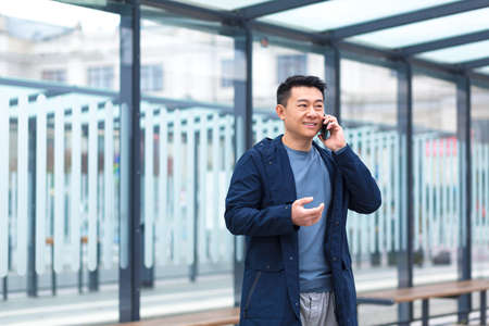 Cheerful and happy Asian businessman talking on the phone, waiting for a plane and a taxi at a public transport stopの写真素材