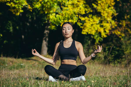 Young beautiful asian woman performs fitness yoga exercises, sitting in lotus position on the grass and meditating in the parkの写真素材