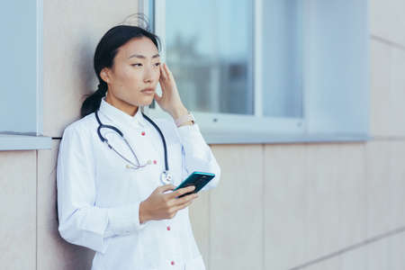 Sad doctor nurse Asian, near the clinic during a break, uses the phone, the woman has a severe headache, depressed overworkerの写真素材