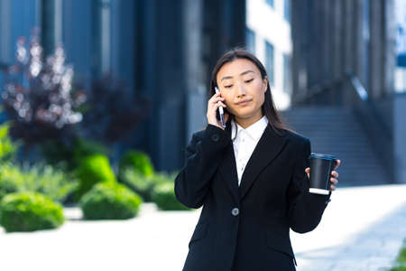 self-possessed asian girl talking on the phone, business woman near the office center on a break, holding a phone and a cup of hot drinkの写真素材