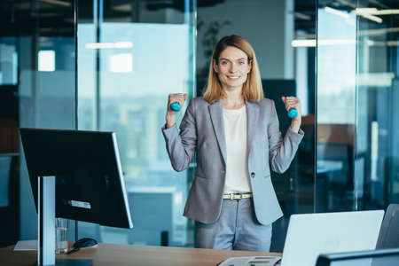 Beautiful and happy woman doing sports exercises in the office at work, during a break, business woman monitors health engaged in fitnessの写真素材