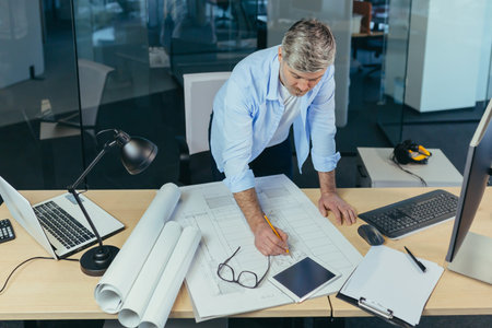 Experienced architect, working at the office desk on the drawing, makes final edits before handing...