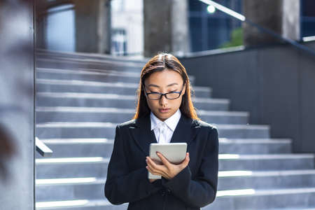 Asian businesswoman in business wear suit standing working with tablet in hands on background modern office building center outside. Male using smartphone, uses mobile phone outdoor city streetの写真素材
