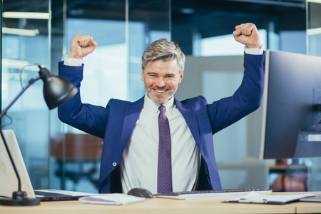 Successful boss works in a modern office at the computer, a senior man gray and happy with a beard...