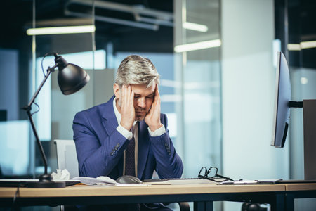 An experienced gray-haired businessman works at the computer until late, tired has a severe headache, stress and depressionの写真素材