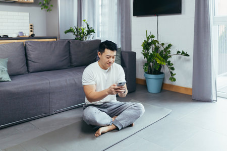 Male asian athlete at home using phone sitting on the floor during morning exercise, athlete in living roomの写真素材