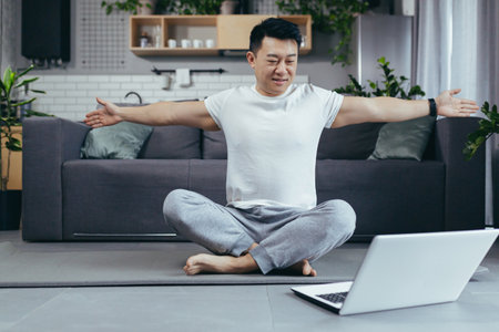 Asian man at home, engaged in fitness online, uses laptop for joint activities, active lifestyleの写真素材