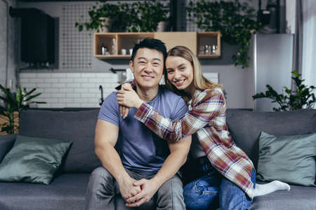 Portrait of young family, man and woman embracing having fun looking at camera while sitting at home on sofaの写真素材