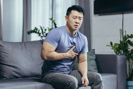A man at home sitting on a sofa in the living room, an Asian patient has severe chest pain, heart diseaseの写真素材