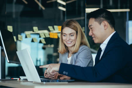 happy coworkers discuss project strategy by looking and pointing at laptop pc computer monitor screen. multiethnic business team in the office. confident mature asian man explaining young femaleの写真素材