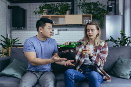 Alcoholic woman drinking hard alcohol at home, husband frustrated and annoyed, conflict in young family over alcoholの写真素材