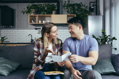 Young family man and woman, multiracial couple sitting on sofa at home, received a happy positive letter with good newsの写真素材