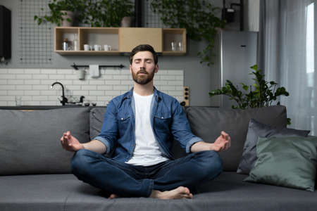 Young man with a beard alone at home, sitting on the couch and meditating in the lotus position, in casual clothesの写真素材