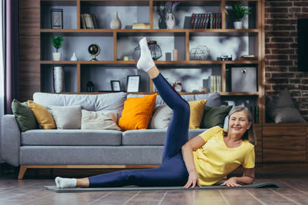 Senior gray-haired woman doing fitness at home, doing morning exercises on a sports mat sitting on the floor at homeの写真素材