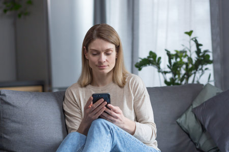 Beautiful middle-aged woman sitting on the couch at home, using the phone and smiling