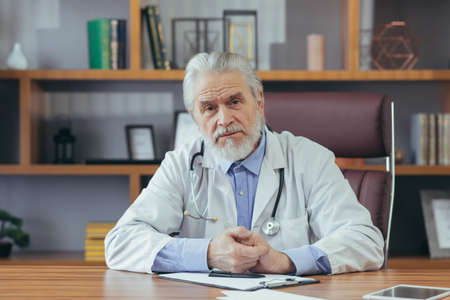 Portrait of a senior experienced doctor gray-haired man working in the office, looking at the camera talking on a video callの写真素材