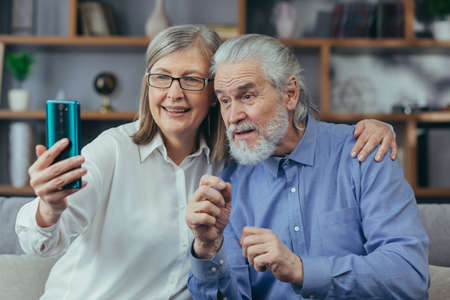 Senior gray-haired married couple man and woman at home together, play video call, sit on sofa at home, use phone for call and online meetingの写真素材