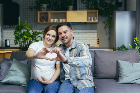 Young couple in love man and pregnant woman sitting on sofa at home and smiling, looking at camera,...