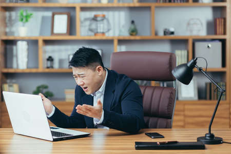 Frustrated and angry asian businessman working on laptop in office, boss shouting at laptop screenの写真素材