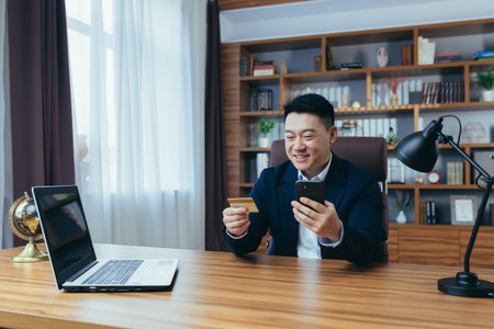 Happy Asian shopping online sitting in the office, man using phone app and bank card, businessman...