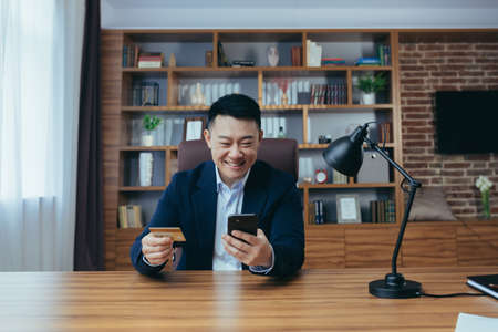 Happy Asian shopping online sitting in the office, man using phone app and bank card, businessman enjoying online shoppingの写真素材