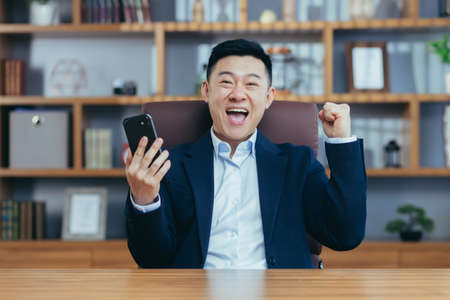 Young happy asian man rejoices won bet, prize, competition, uses phone. In the office, sitting at a table in a suitの写真素材