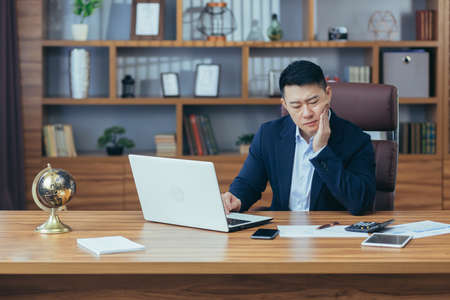 Toothache in the workplace. A young Asian man, a businessman, an employee, has a toothache at work. He is sitting at a desk in the office, holding his cheek, feeling painの写真素材