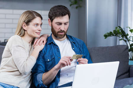Happy married couple man and woman sitting at home on the couch using laptop for online shopping, holding a credit bank cardの写真素材